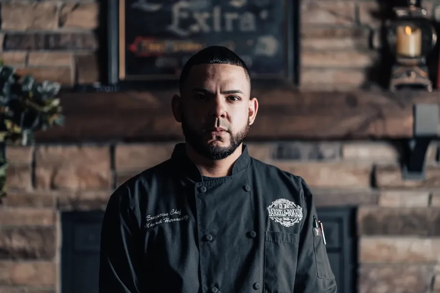 Chef Manny Hernandez in his black chef jacket beside a stone fireplace