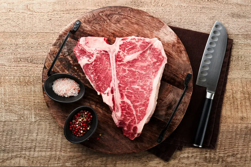 A raw porterhouse on a wooden board with sea salt and cracked pepper
