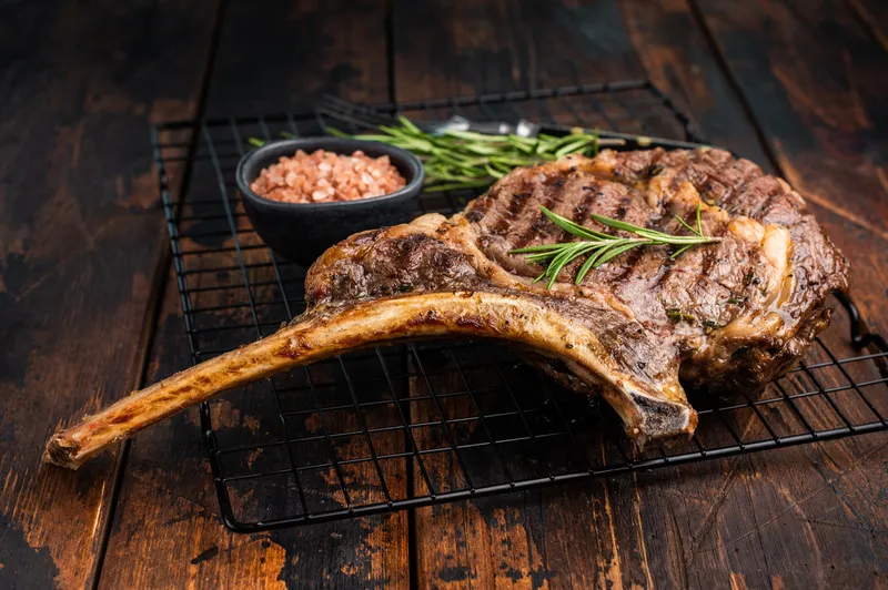A bone-in tomahawk ribeye resting on a cooling rack with rosemary and salt